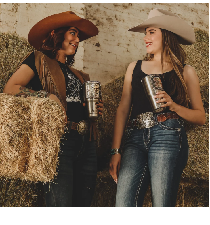 Two women in cowboy hats on hay bales wearing engraved Classic belt buckles