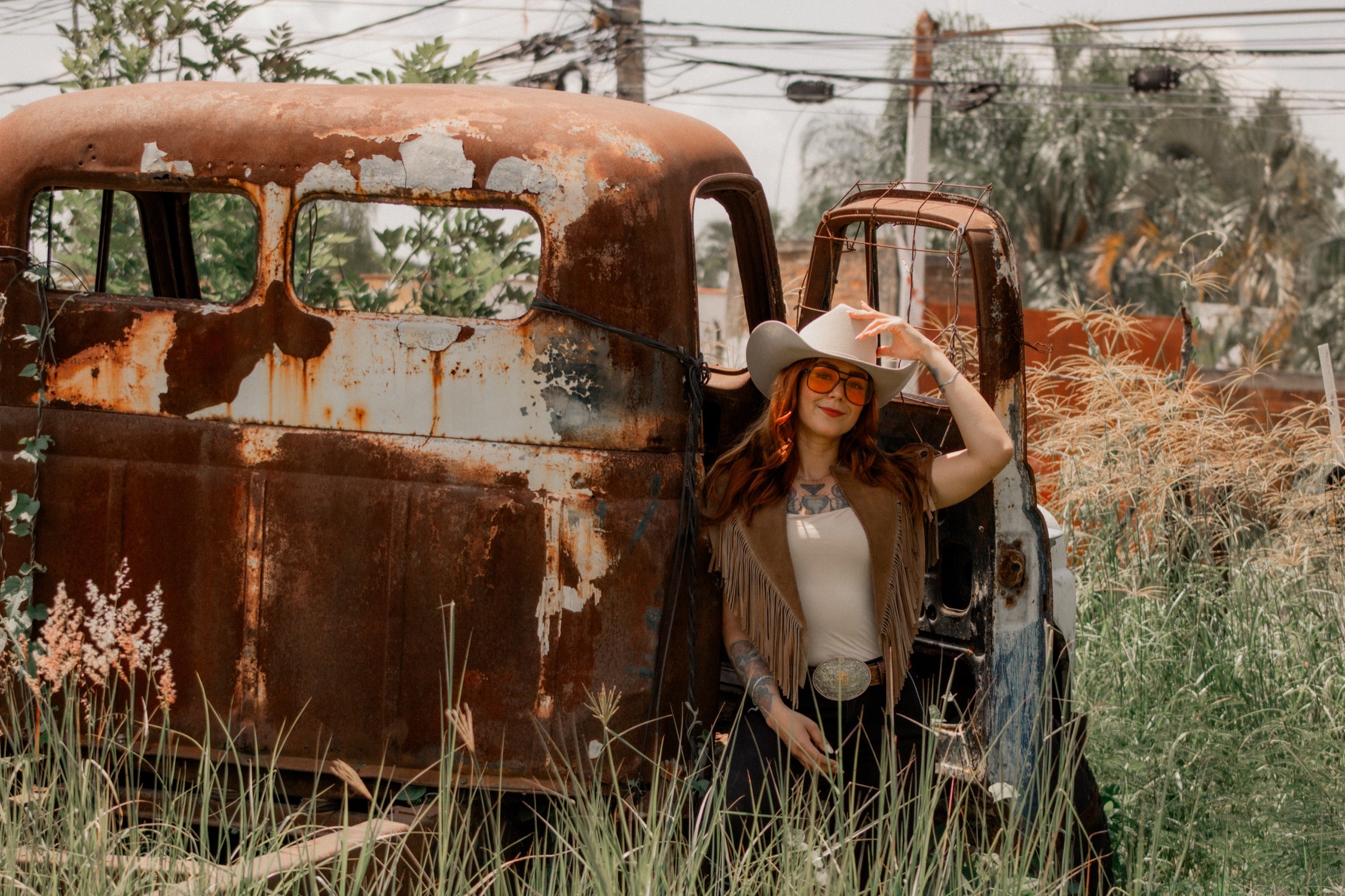 Woman in a fringe vest and cowboy hat wearing an engraved trophy buckle beside a rusted vintage truck