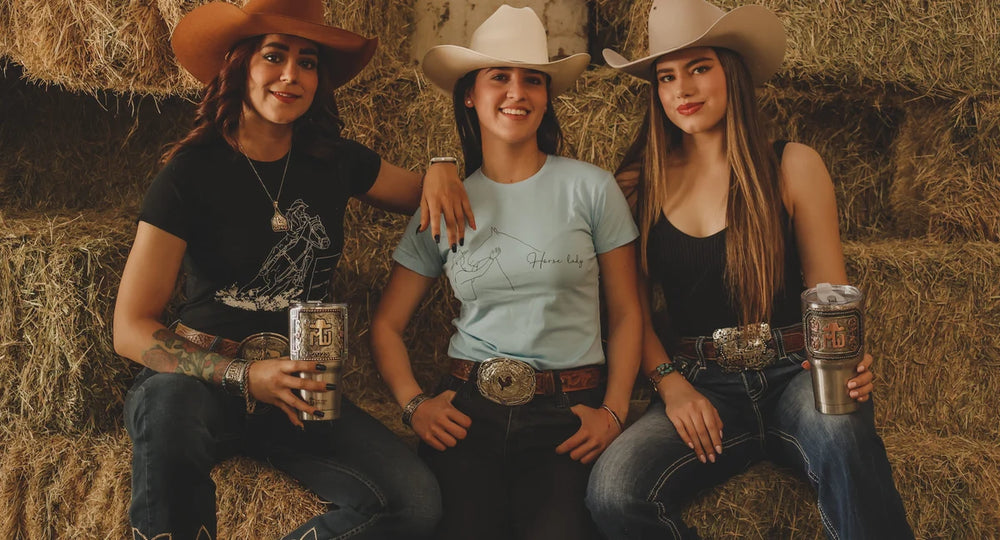 Three women in cowboy hats on hay bales wearing engraved trophy buckles and holding matching engraved tumblers