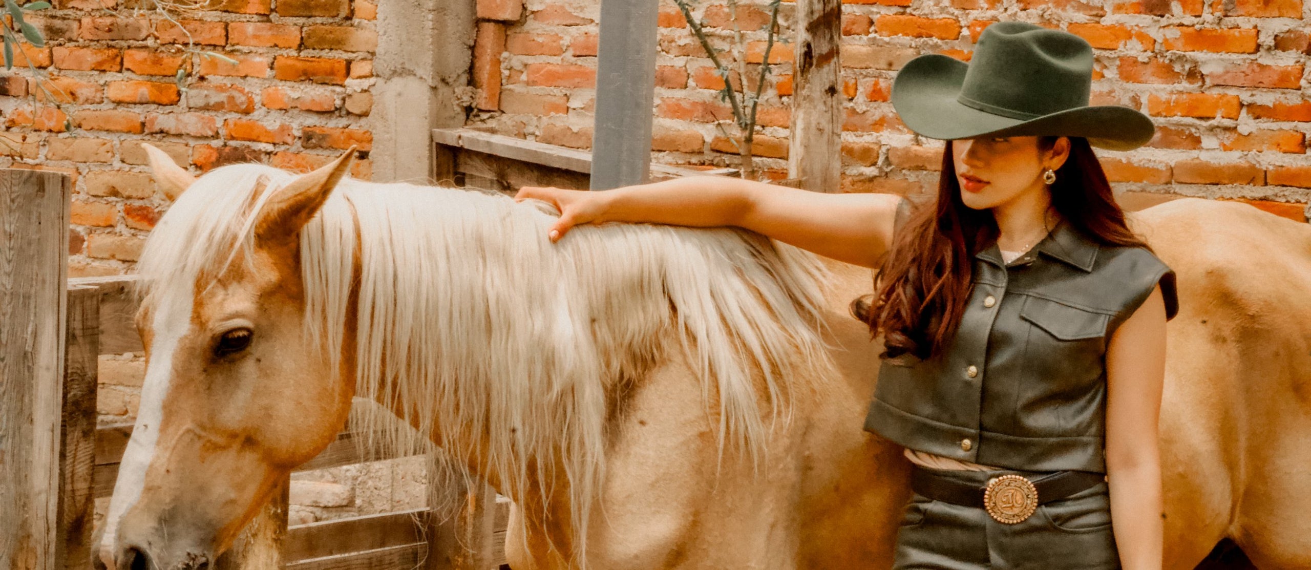 Woman in a green cowboy hat and leather set wearing a Most Wanted engraved buckle beside a palomino horse